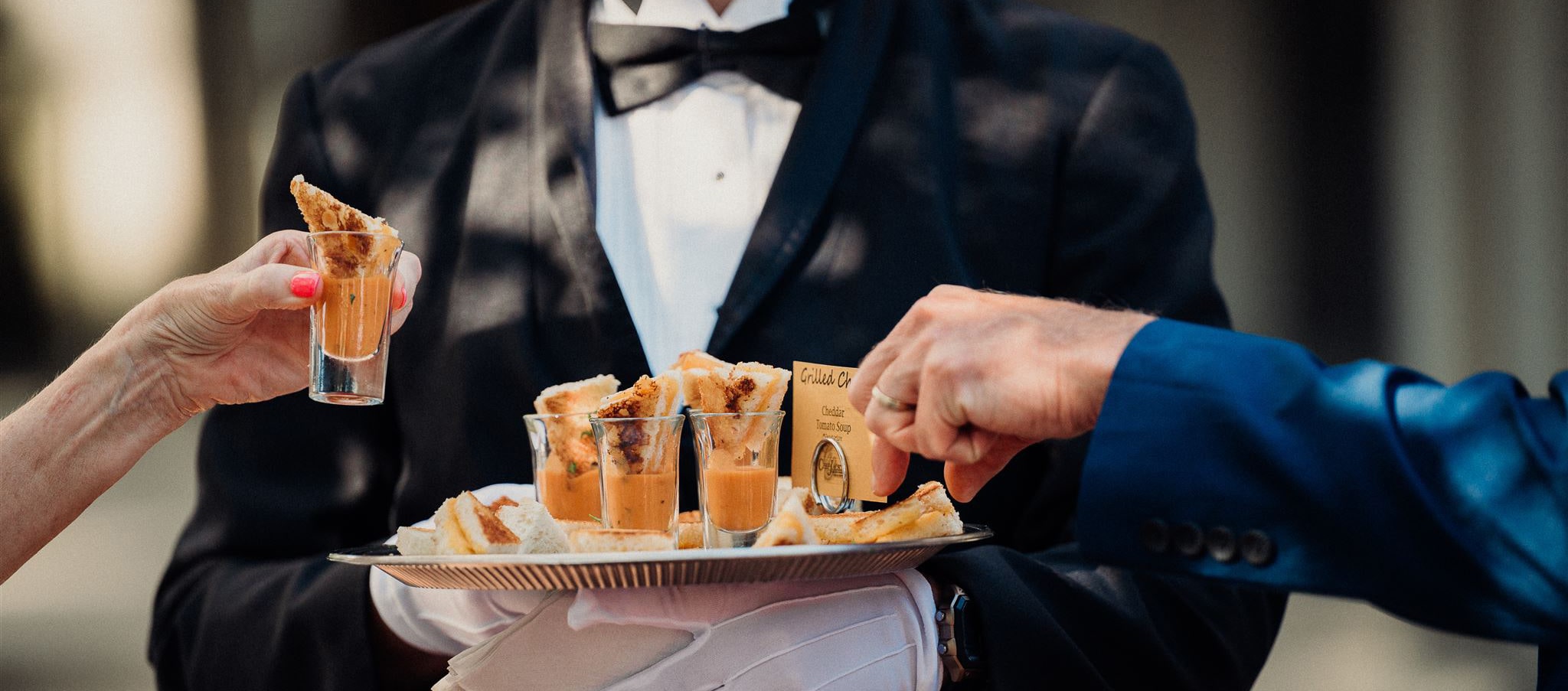 A waiter in a tuxedo holding a plate of grilled cheese sandwich bite in shot glasses with cheese at the bottom appetizers. Two people are grabbing an appetizer.