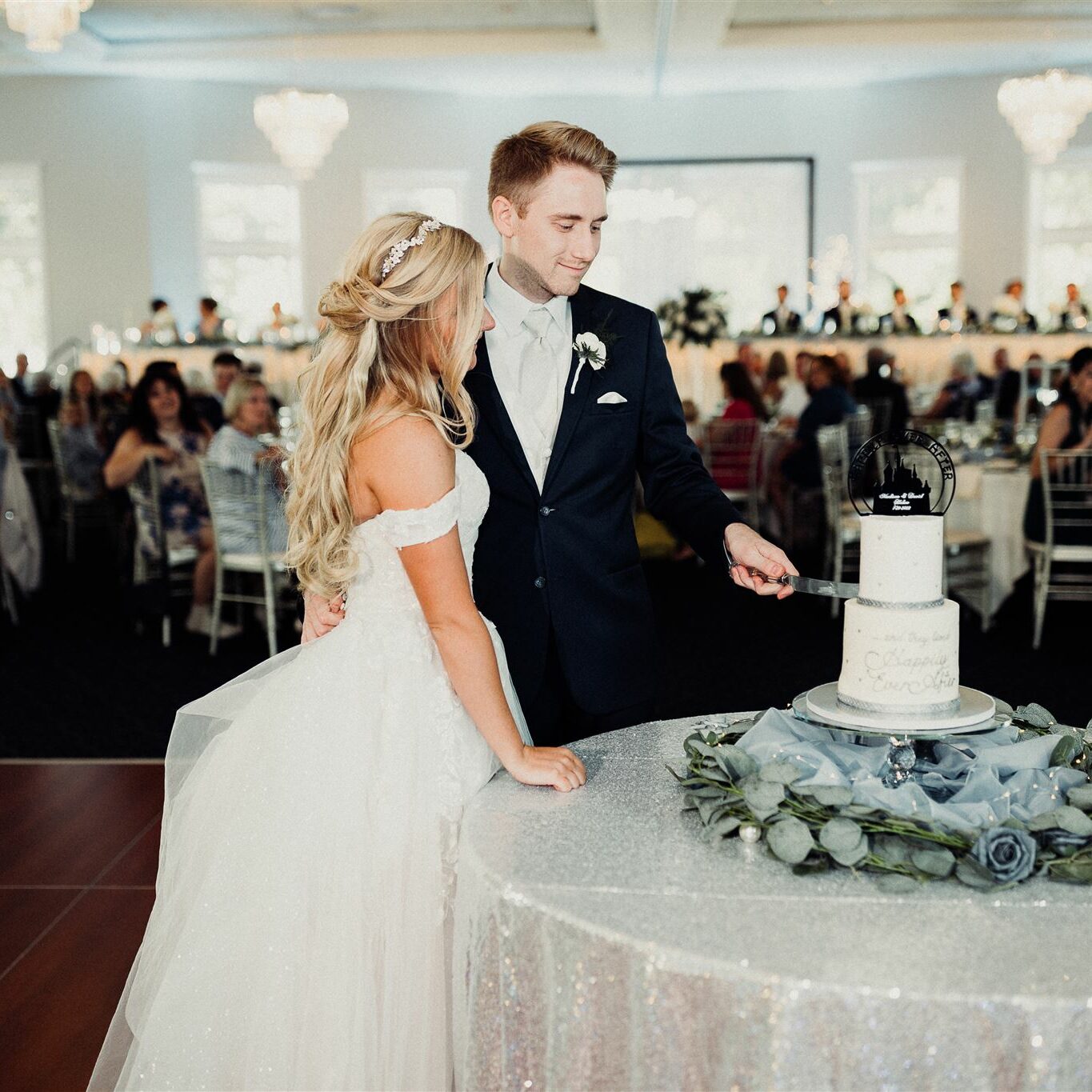 A bride and groom cutting a two layer wedding cake while the guests watch from their tables in the background.